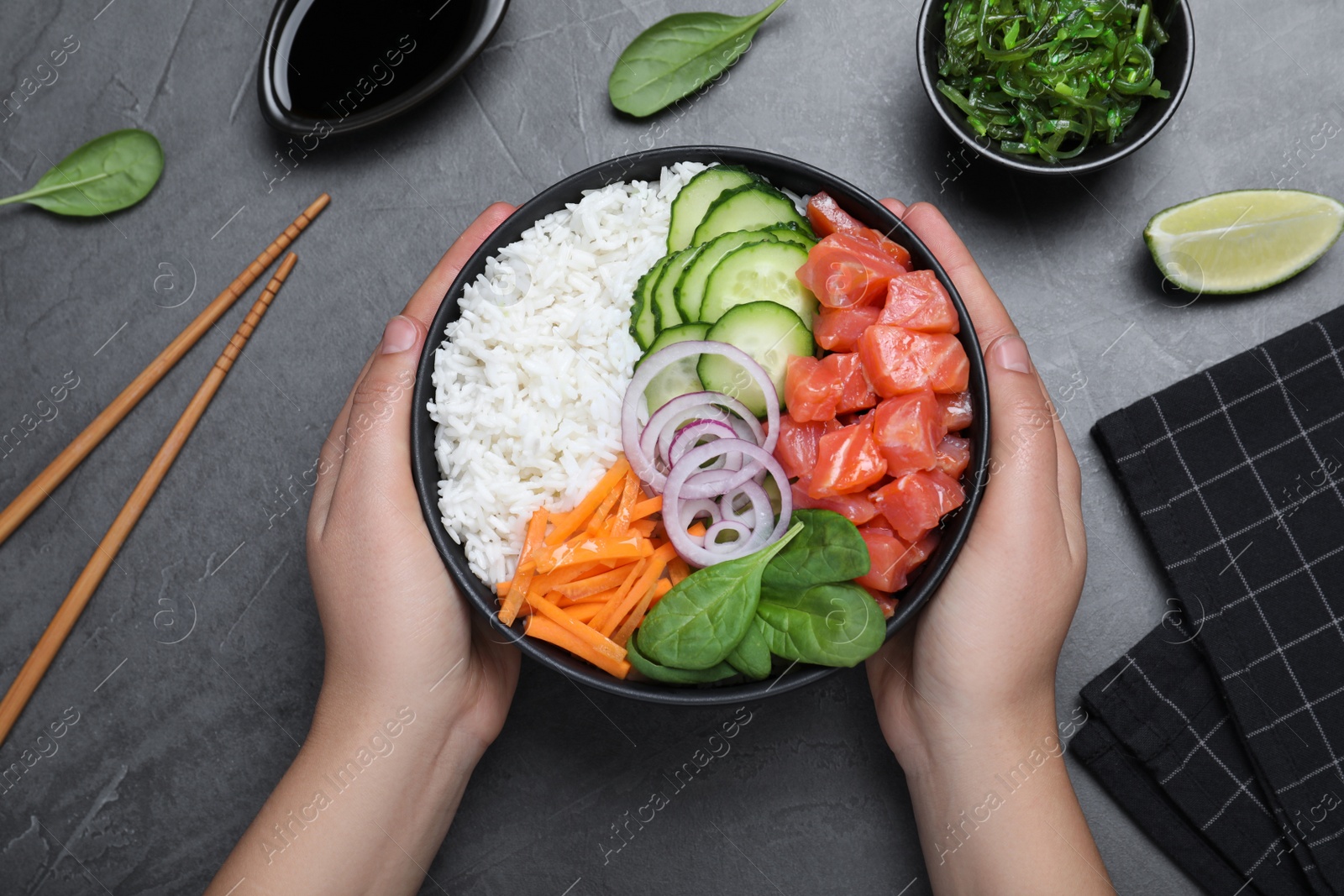 Photo of Woman holding delicious poke bowl with salmon and vegetables at grey table, top view