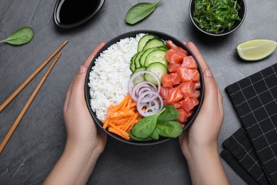 Photo of Woman holding delicious poke bowl with salmon and vegetables at grey table, top view