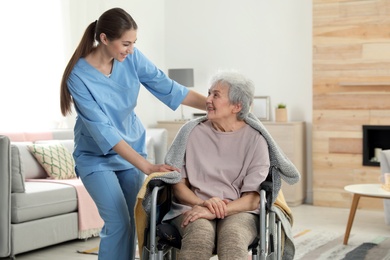 Photo of Nurse covering elderly woman in wheelchair with blanket indoors. Assisting senior people