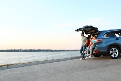 Photo of Happy young family sitting in car trunk on riverside