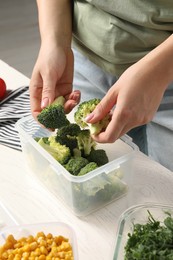 Woman putting broccoli into plastic container at white wooden table, closeup. Food storage