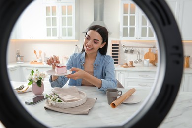Photo of Blogger with cake recording video in kitchen at home, view through ring lamp