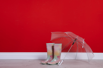 Photo of Beautiful transparent umbrella and rubber boots near red wall. Space for text