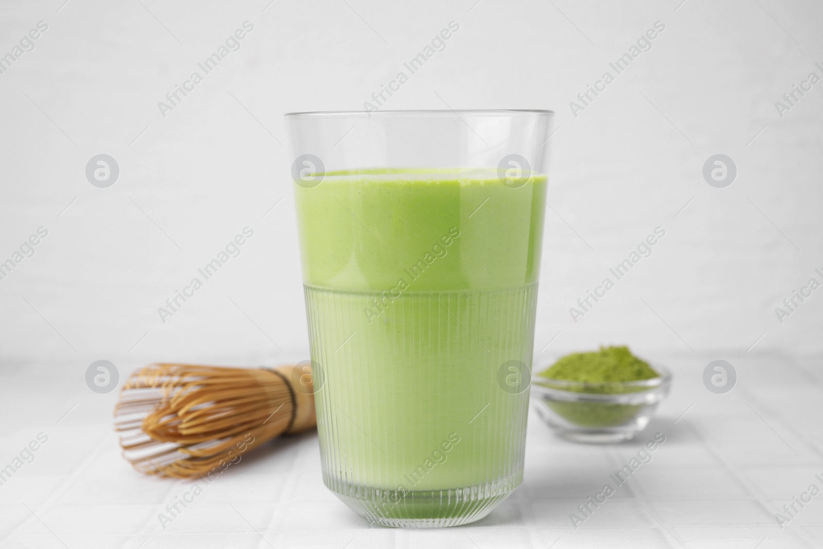 Photo of Glass of tasty matcha smoothie, powder and bamboo whisk on white tiled table, closeup