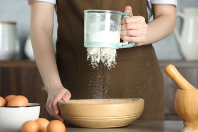 Woman sieving flour into bowl at table in kitchen, closeup