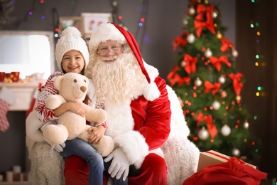 Little girl with teddy bear sitting on authentic Santa Claus' lap indoors