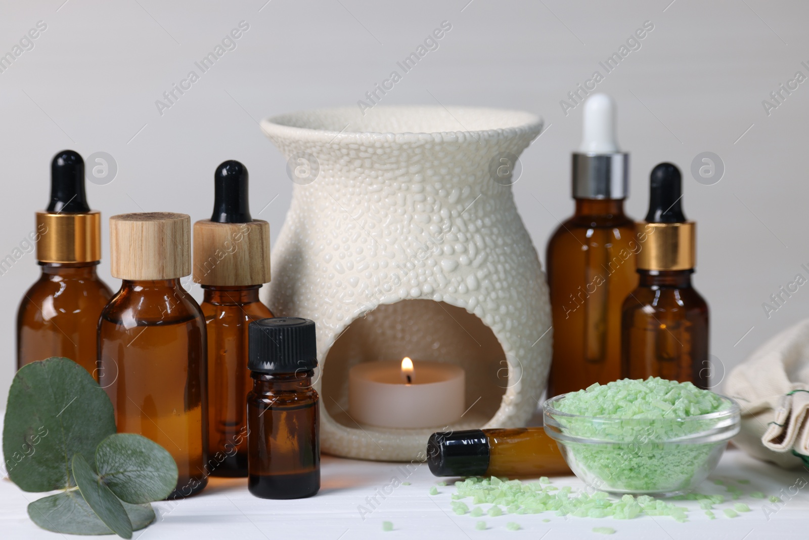 Photo of Different aromatherapy products, burning candle and eucalyptus leaves on white wooden table against light background, closeup