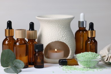 Different aromatherapy products, burning candle and eucalyptus leaves on white wooden table against light background, closeup