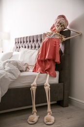 Photo of Human skeleton in silk pajamas and towel sitting on bed indoors