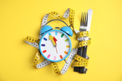 Photo of Alarm clock, measuring tape and cutlery on yellow background, flat lay. Diet regime