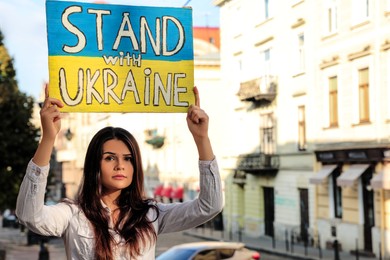 Photo of Sad woman holding poster in colors of national flag and words Stand with Ukraine on city street. Space for text