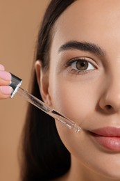 Beautiful young woman applying serum onto her face on beige background, closeup