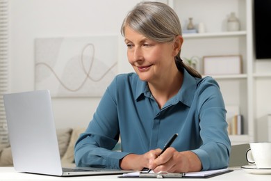 Photo of Beautiful senior woman taking notes while working with laptop at white table indoors