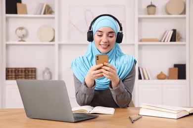 Muslim woman using smartphone near laptop at wooden table in room