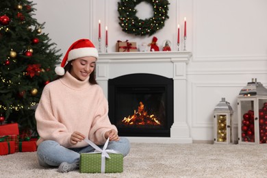 Happy young woman in Santa hat opening gift box in room decorated for Christmas