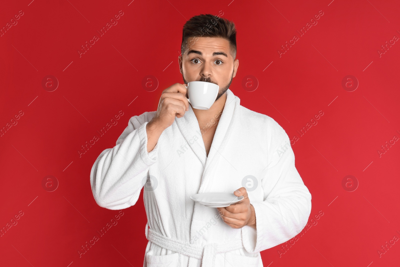 Photo of Young man in bathrobe with cup of coffee on red background