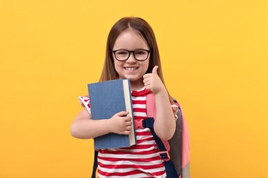 Photo of Cute little girl with backpack and book showing thumbs up against orange background