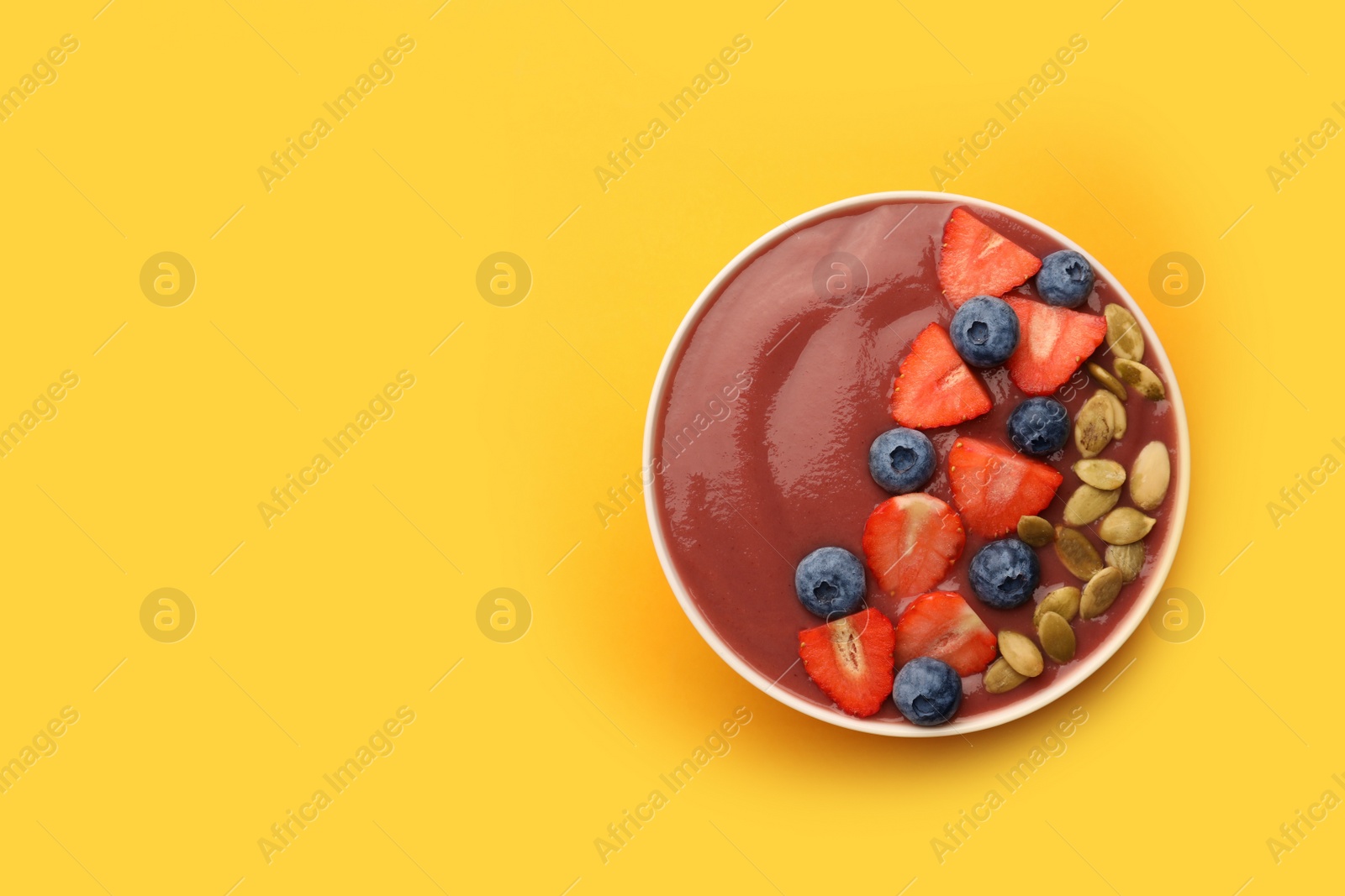 Photo of Bowl of delicious smoothie with fresh blueberries, strawberries and pumpkin seeds on yellow background, top view. Space for text