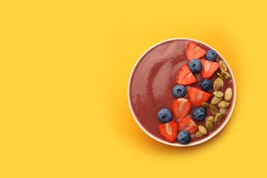 Bowl of delicious smoothie with fresh blueberries, strawberries and pumpkin seeds on yellow background, top view. Space for text