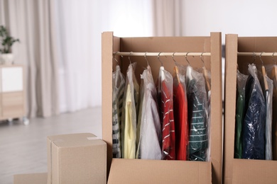 Photo of Cardboard wardrobe boxes with clothes on hangers indoors. Moving day