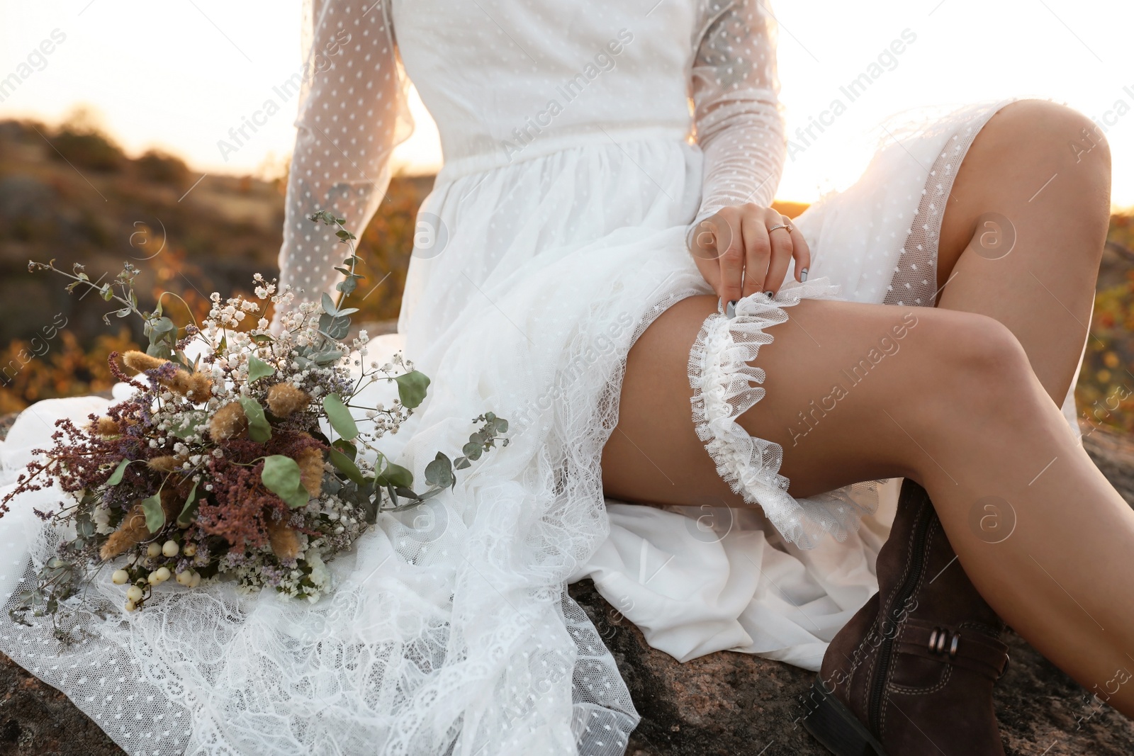 Photo of Beautiful bride with field bouquet sitting on rock at sunset, closeup