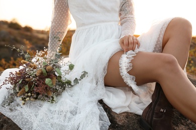 Photo of Beautiful bride with field bouquet sitting on rock at sunset, closeup