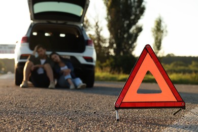 Photo of Warning triangle and couple sitting near broken car on roadside, selective focus