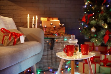 Photo of Cups and jar with cookies on table in room decorated for Christmas. Stylish interior