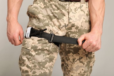 Soldier in military uniform applying medical tourniquet on leg against light grey background, closeup