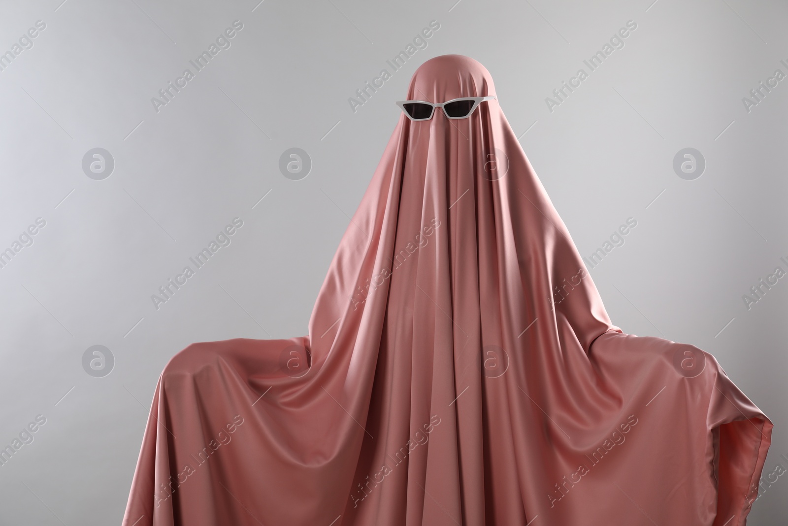 Photo of Glamorous ghost. Woman in pink sheet with sunglasses on light grey background, space for text