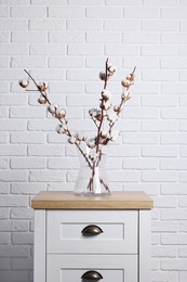 Cotton branches with fluffy flowers in vase on wooden commode near white brick wall