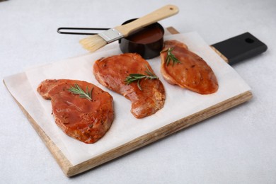 Photo of Board with raw marinated meat, rosemary and basting brush on light table, closeup