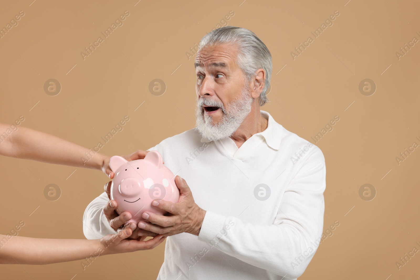 Photo of Woman taking piggy bank from confused senior man on beige background. Be careful - fraud