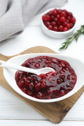 Photo of Tasty cranberry sauce in bowl, fresh berries and rosemary on white wooden table, closeup