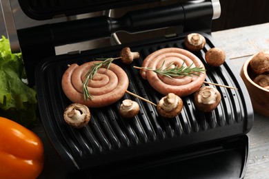 Photo of Electric grill with homemade sausages, rosemary and mushrooms on rustic wooden table