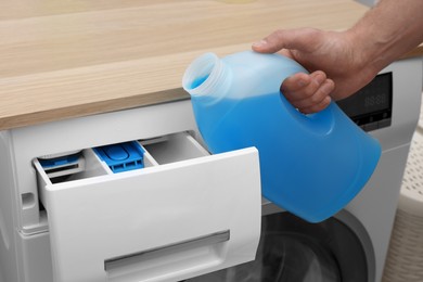 Photo of Man pouring fabric softener from bottle into washing machine indoors, closeup