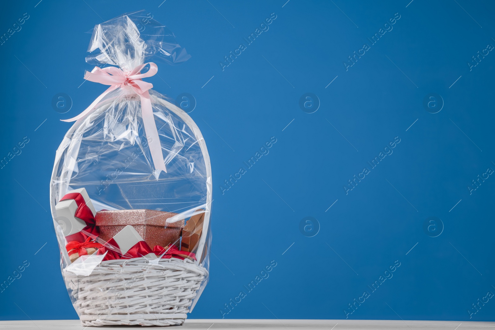 Photo of Wicker basket full of gift boxes on white wooden table against blue background. Space for text
