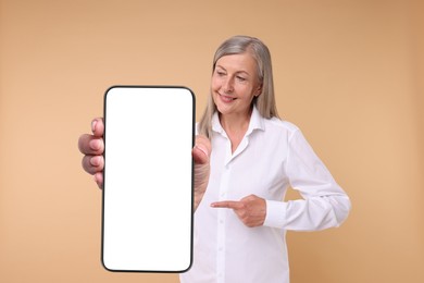 Happy mature woman pointing at mobile phone with blank screen on beige background. Mockup for design