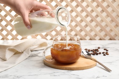 Photo of Woman pouring milk into glass with refreshing iced coffee at white marble table, closeup