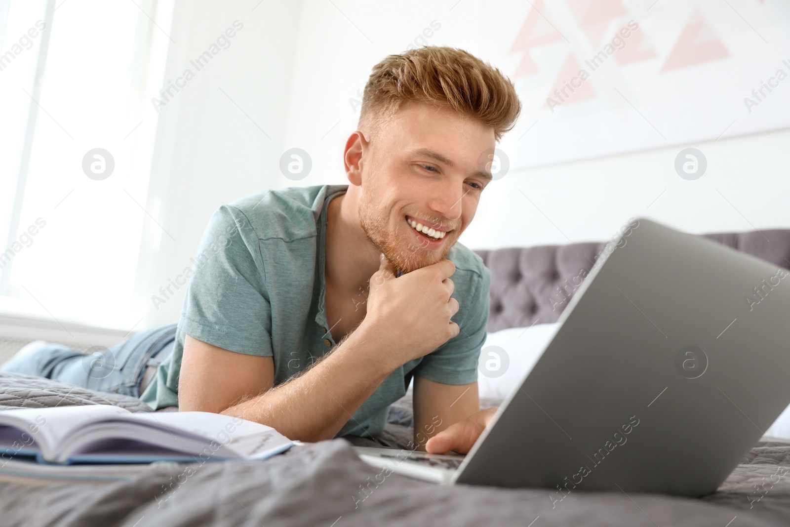 Photo of Young man using laptop while lying on bed at home