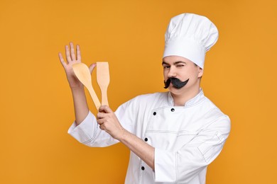 Photo of Portrait of happy confectioner with funny artificial moustache holding wooden spatulas on orange background