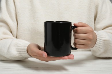 Photo of Woman holding black mug at white table, closeup. Mockup for design