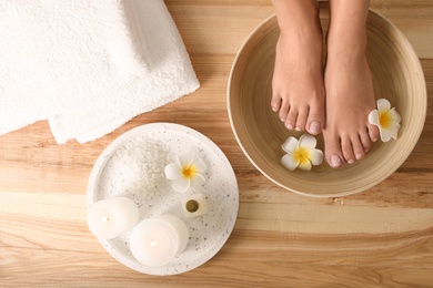 Woman soaking her feet in dish with water and flowers on wooden floor, top view. Spa treatment