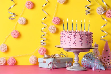 Photo of Delicious birthday cake and party decor on pink table against yellow background