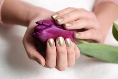 Woman with gold manicure holding flower on light background, closeup. Nail polish trends