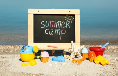 Photo of Small chalkboard with text SUMMER CAMP and beach toys on sand near river