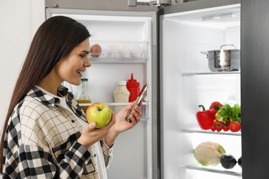 Young woman with apple and smartphone near modern refrigerator indoors