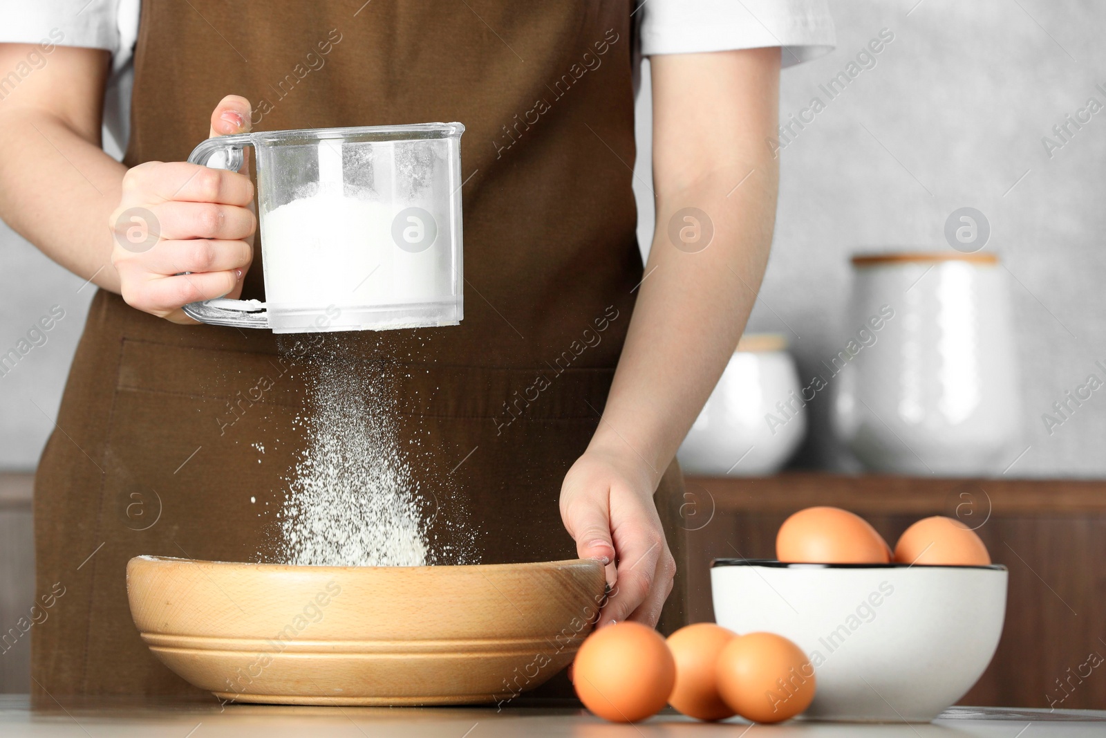 Photo of Woman sieving flour into bowl at table in kitchen, closeup