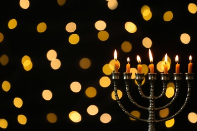 Photo of Golden menorah with burning candles against dark background and blurred festive lights, space for text