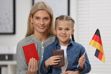Photo of Immigration. Happy woman with her daughter holding passports and flag of Germany indoors